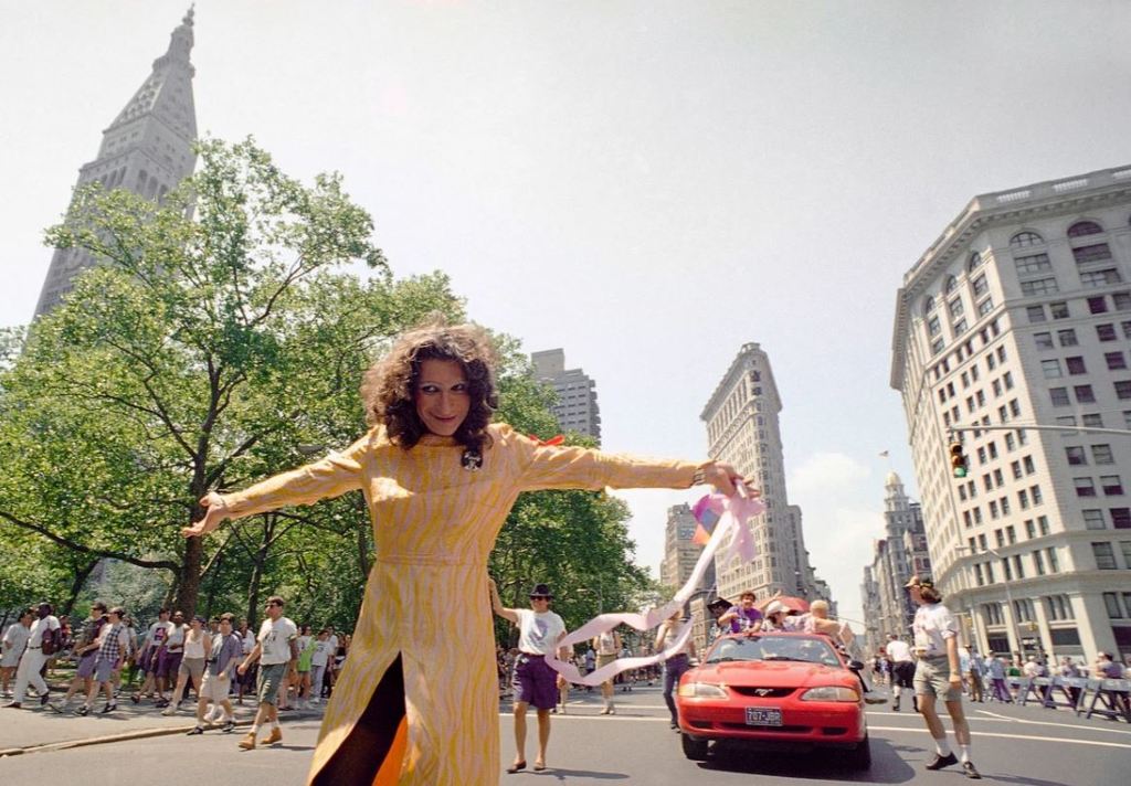 Image of Sylvia Rivera in a yellow dress and black tights in a street with people in the background. Summer day