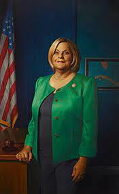 Photo of Lleana ros-lehtinen in a blue dress and green coat standing next to a flag in front of a dark blue wall